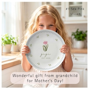 This cream-colored plate features a "Sea Pink" botanical design. At the center is a cluster of soft pink flowers on green stems, with the name "gigi" written in dark green cursive and "est 2020" in lowercase block text below. Two decorative rings frame the center: an inner light blue scalloped lace border and an outer sage green vine border. Small, individual watercolor leaves are scattered between the rings, creating a delicate, garden-inspired look.  Wonderful gift for Grandma on Mother's Day!