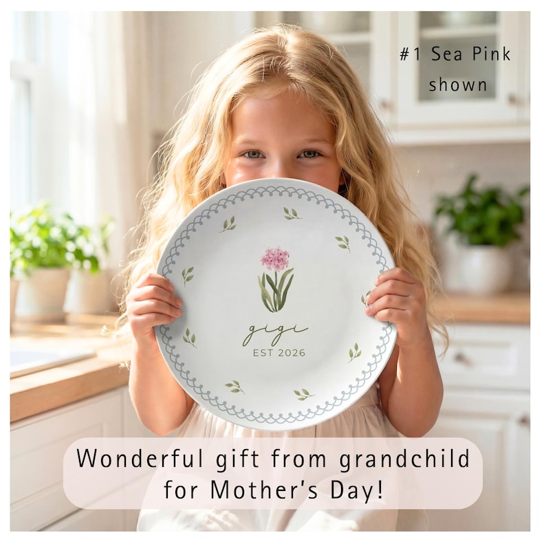 This cream-colored plate features a "Sea Pink" botanical design. At the center is a cluster of soft pink flowers on green stems, with the name "gigi" written in dark green cursive and "est 2020" in lowercase block text below. Two decorative rings frame the center: an inner light blue scalloped lace border and an outer sage green vine border. Small, individual watercolor leaves are scattered between the rings, creating a delicate, garden-inspired look.  Wonderful gift for Grandma on Mother's Day!
