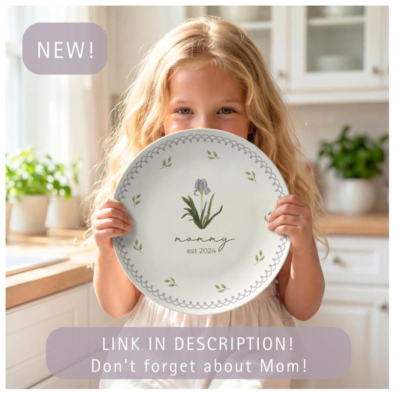 May include: A white plate with a floral design, held by a child. The plate features a purple iris, green leaves, and the word "mommy est. 2024". The rim has a scalloped edge with small green leaf accents. The word "NEW!" is in the upper left corner.
