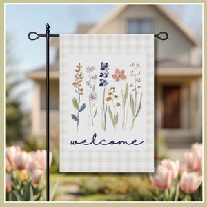 Image of garden flag, featuring a soft watercolor illustration of delicate wildflowers in shades of lavender, peach, and blue. Set against a classic light beige gingham border, the elegant navy script reads "welcome," creating a warm invitation for guests. This durable, double-sided flag is perfect for spring and summer decor, adding a touch of cottagecore aesthetic to your flower beds or walkway.