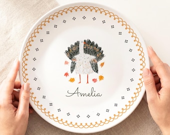 May include: A white ceramic plate featuring a central illustration of a turkey with dark gray and white feathers, surrounded by small orange and red autumn leaves. The name Amelia is printed in black script below the turkey, with a gold scalloped border.