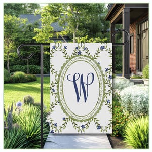 Puede incluir: Una bandera de jardín blanca con la letra "W" azul marino dentro de un marco ovalado, rodeada de un diseño floral verde y flores azules. La bandera se muestra en un soporte de metal negro en un jardín.