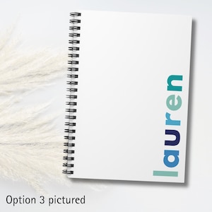 A white spiral bound notebook with the name 'lauren' printed on the cover in blue, teal, and green letters.
