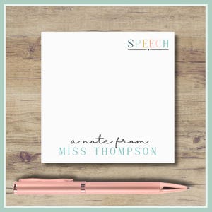 May include: A white notepad with the word "SPEECH" in teal and black, and the phrase "a note from MISS THOMPSON" in teal script. A rose gold pen rests below the notepad on a wooden surface.