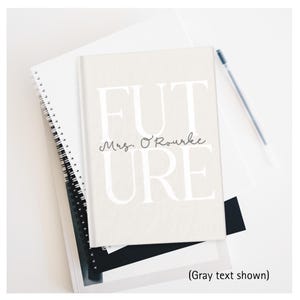 May include: A light beige notebook with the word "FUTURE" in large white letters and "Mrs. O'Rourke" in cursive. The notebook is on top of other notebooks and a pen.