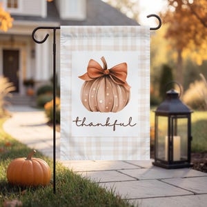 Può includere: Una bandiera da giardino rettangolare con un motivo a quadri beige chiaro e bianco. Il centro mostra un'illustrazione ad acquerello di una zucca color marrone chiaro con pois bianchi e un grande fiocco arancione, con la scritta thankful in marrone.