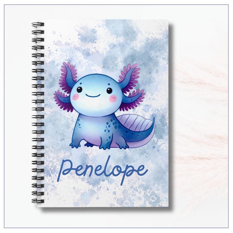 Image of the notebook cover.  A cheerful cartoon axolotl with a round, smiling face and rosy cheeks. Its body is light blue with darker blue spots and shading, and it has large frilly gills on each side of its head in shades of purple and pink. The background is a soft watercolor-style wash of blues and whites, giving a dreamy, splashy effect. At the bottom, the name “Penelope” is written in playful, dark blue script lettering. The notebook is spiral-bound along the left edge.