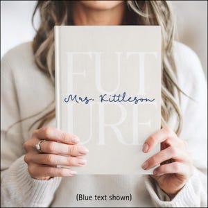 A woman is holding a light beige hardcover journal in front of her chest with both hands. The journal cover has the large word “FUTURE” printed in a soft white font across the front. The letters F, U, and T are on the top row with the letters U, R, and E below. Across the middle, the name “Mrs. Kittleson” is written in a dark blue cursive script. The person holding the journal is wearing a white sweater, and the background is softly blurred.