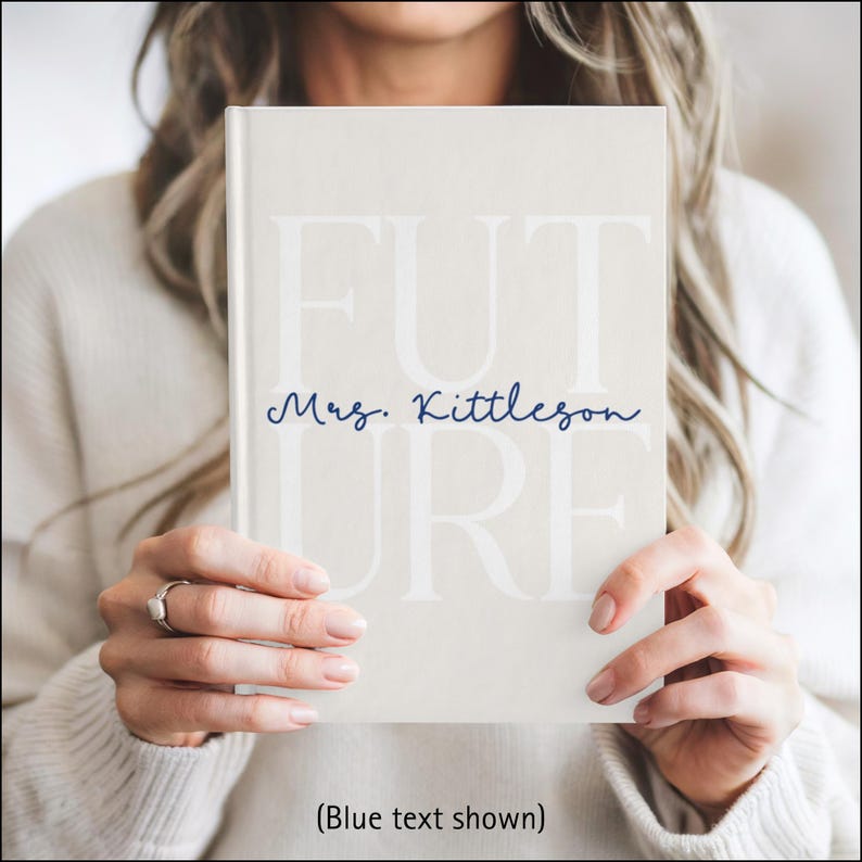 A woman is holding a light beige hardcover journal in front of her chest with both hands. The journal cover has the large word “FUTURE” printed in a soft white font across the front. The letters F, U, and T are on the top row with the letters U, R, and E below. Across the middle, the name “Mrs. Kittleson” is written in a dark blue cursive script. The person holding the journal is wearing a white sweater, and the background is softly blurred.