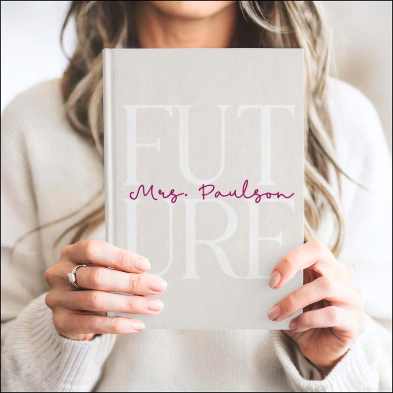 A woman is holding a light beige hardcover journal in front of her chest with both hands. The journal cover has the large word “FUTURE” printed in a soft white font across the front. The letters F, U, and T are on the top row with the letters U, R, and E below. Across the middle, the name “Mrs. Kittleson” is written in a raspberry-colored cursive script. The person holding the journal is wearing a white sweater, and the background is softly blurred.