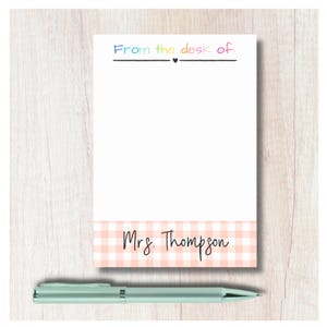 May include: A white notepad with the text "From the desk of." in rainbow colours and "Mrs. Thompson" in black script over a peach gingham pattern. A silver pen rests below the notepad.