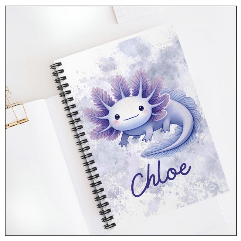 Image of the notebook cover laying on a desk.  A cheerful cartoon axolotl with a round, smiling face and rosy cheeks. Its body is light purple. It has large frilly gills on each side of its head in shades of purple. The background is a soft watercolor-style wash of purples, giving a dreamy, splashy effect. At the bottom, the name “Chloe” is written in playful, dark purple script lettering. The notebook is spiral-bound along the left edge.