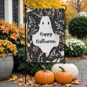 May include: A Halloween garden flag featuring a white ghost with the words "Happy Halloween" on a black background with floral accents. The flag is displayed outdoors with orange pumpkins and fall foliage.
