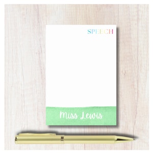 May include: A white notepad with the word "SPEECH" in rainbow colors at the top and "Miss Lewis" in white script at the bottom. A gold pen rests below the notepad on a light wood surface.