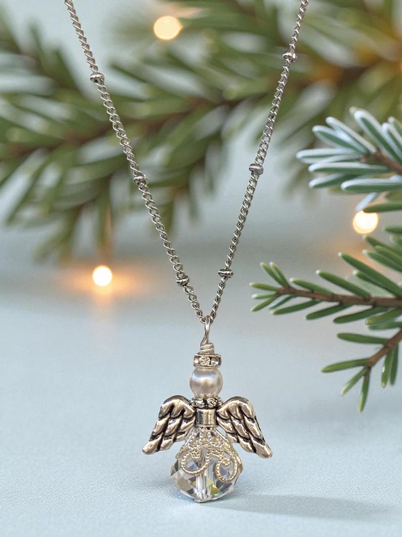 May include: A silver-toned angel pendant necklace. The pendant features an angel with detailed wings and a crystal-like body. The necklace chain is delicate and hangs against a backdrop of evergreen branches and soft, out-of-focus lights.