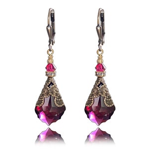 May include: A pair of dangle earrings with a teardrop-shaped purple crystal accented with gold and silver details. The earrings have a clip-on closure.