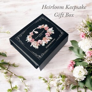 May include: A black gift box with the text "Heirloom Keepsake Gift Box" and a floral design on the lid. The floral design includes pink and white elements. The box is surrounded by flowers.