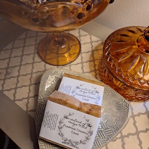 May include: Two bars of pumpkin crunch cake soap on a decorative silver tray. Each bar is wrapped with a label that reads "Homestead of the Hedge Witch PUMPKIN CRUNCH CAKE" and "Made in Dublin, OH." A decorative glass bowl and jar are in the background.