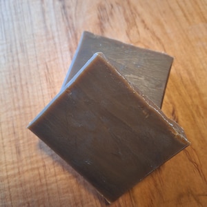 May include: Two rectangular bars of brown soap are stacked on a wooden surface. The soap has a smooth, matte finish and a slightly irregular shape. The bars are angled towards each other, creating a sense of depth.