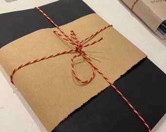 Puede incluir: Cuadernos negros envueltos en papel marrón y atados con hilo rojo y dorado. Se ven más cuadernos en el fondo, algunos envueltos en papel azul. Los cuadernos están sobre una superficie blanca.