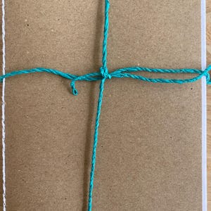 Pode incluir: Um caderno de cartão castanho com um cordão turquesa amarrado em volta em forma de cruz.