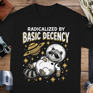 以下が含まれることがあります： 「RADICALIZED BY BASIC DECENCY」の文字が入った黒いTシャツ。宇宙服を着た漫画のキャラクターのアライグマが、輪のある惑星と星の近くを浮遊しているデザインです。面白くて楽しいデザインです。