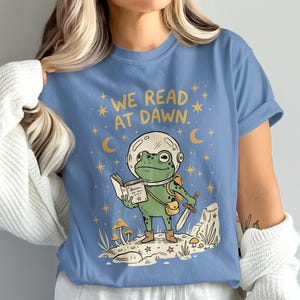 Peut inclure: T-shirt bleu clair avec une illustration d'une grenouille en casque spatial lisant un livre et tenant une épée. Le texte "WE READ AT DAWN" est au-dessus de la grenouille, avec des étoiles et des croissants de lune.