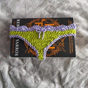 May include: A handmade crochet bikini bottom featuring a vibrant lime green center panel with a scalloped lavender trim along the waistband and leg openings. A small lavender crochet bow is centered at the front waistband of the garment.