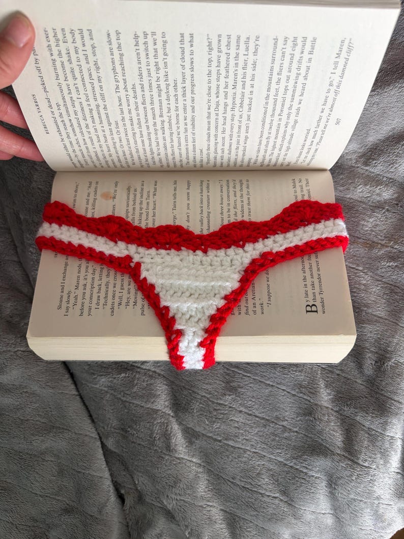 May include: A handmade crochet thong featuring a horizontal striped pattern in vibrant red and white yarn. The garment has a scalloped edge along the top waistband and a narrow, tapered design that extends downward, resting on an open book.