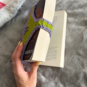 May include: A lime green and lavender crochet book sleeve featuring a textured stitch pattern and a lavender border. The sleeve is wrapped around a paperback book with a black cover that displays the text YARROS in white capital letters.