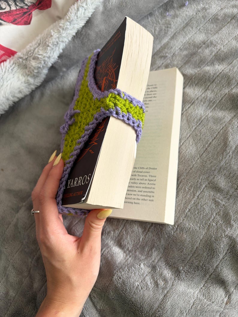 May include: A lime green and lavender crochet book sleeve featuring a textured stitch pattern and a lavender border. The sleeve is wrapped around a paperback book with a black cover that displays the text YARROS in white capital letters.