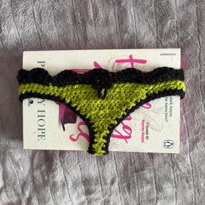 May include: A crochet thong featuring a lime green center panel with a black scalloped border and trim. The item is positioned over a book titled 'The Mystery of Mercy Close' by Marian Keyes, which has white covers and pink text.