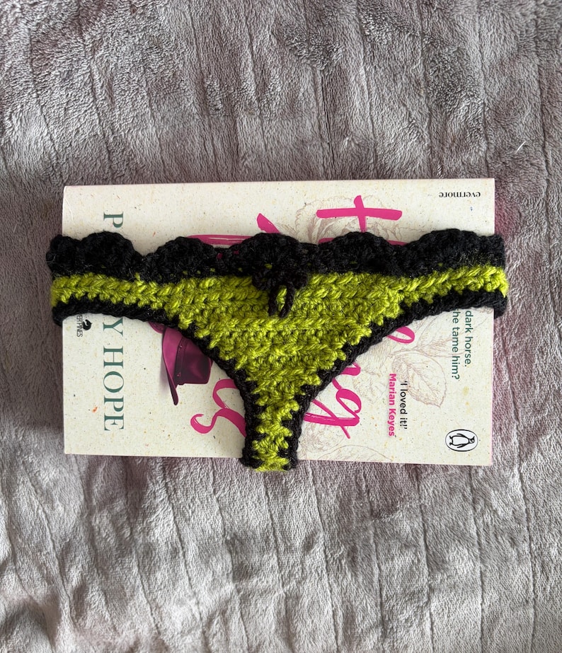 May include: A crochet thong featuring a lime green center panel with a black scalloped border and trim. The item is positioned over a book titled 'The Mystery of Mercy Close' by Marian Keyes, which has white covers and pink text.