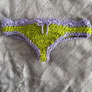 May include: A crochet bikini bottom featuring a lime green center panel and a lavender scalloped waistband. The garment includes a small lavender drawstring tie at the center front and lavender trim along the leg openings, crafted from yarn.
