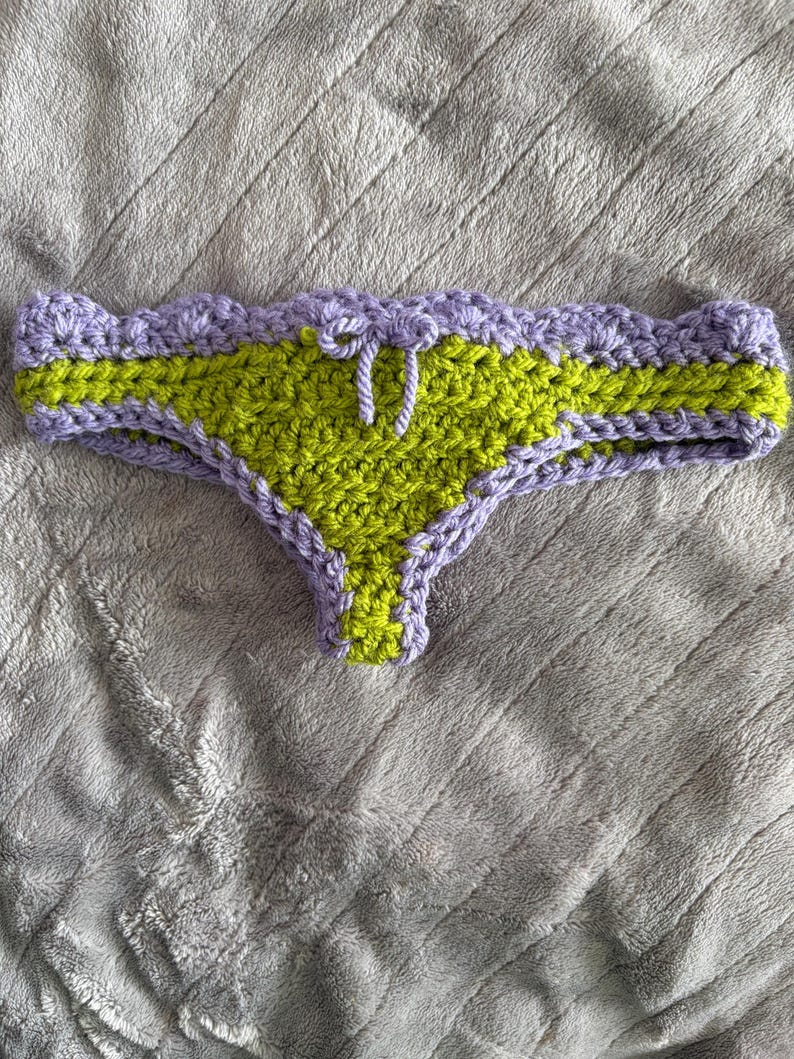 May include: A crochet bikini bottom featuring a lime green center panel and a lavender scalloped waistband. The garment includes a small lavender drawstring tie at the center front and lavender trim along the leg openings, crafted from yarn.