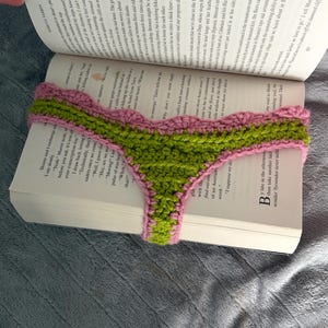 May include: A handmade crochet bookmark featuring a vibrant green center with a textured stitch pattern, bordered by a scalloped edge in a soft pink yarn. The bookmark is draped horizontally across the open pages of a printed hardcover book.