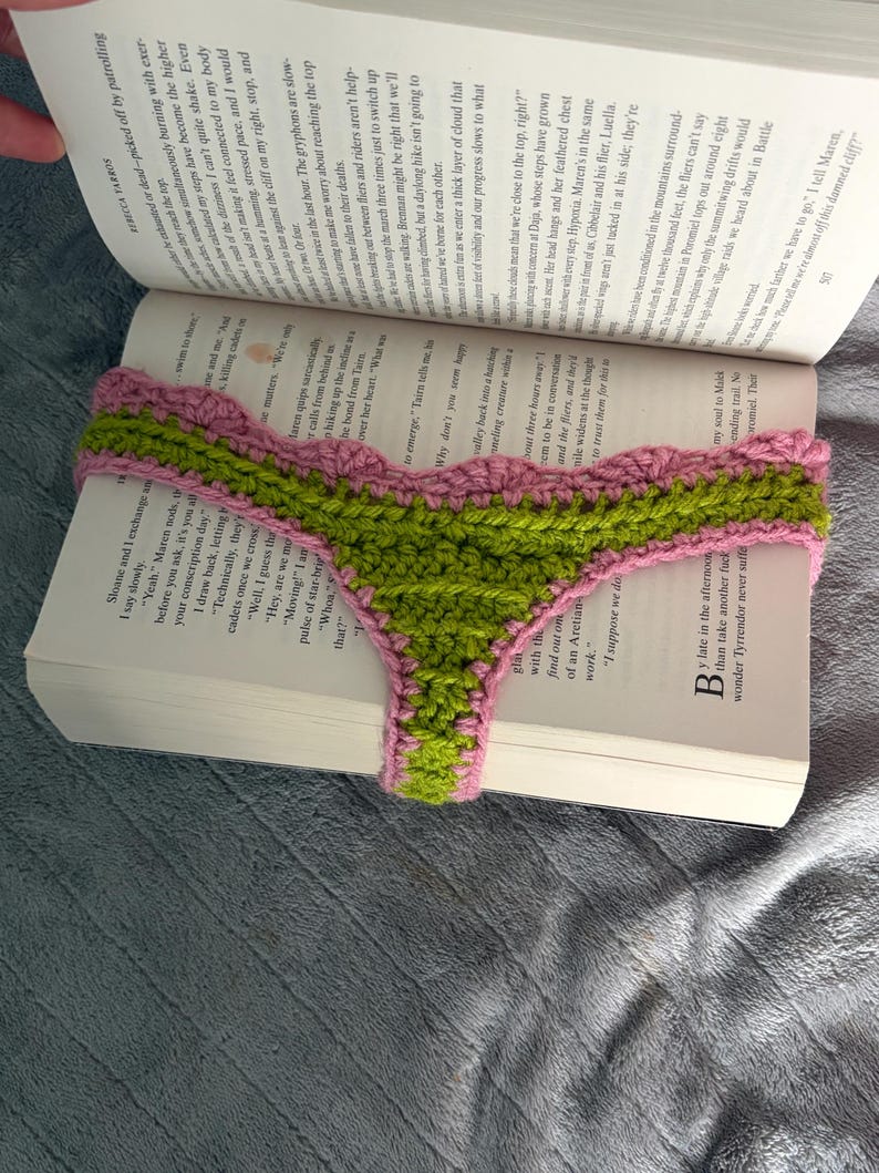 May include: A handmade crochet bookmark featuring a vibrant green center with a textured stitch pattern, bordered by a scalloped edge in a soft pink yarn. The bookmark is draped horizontally across the open pages of a printed hardcover book.