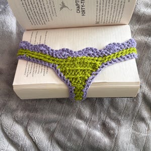 May include: A handmade crochet bookmark featuring a triangular lime green center and a scalloped lavender purple border. The bookmark is draped over the open pages of a book, with the text Chapter Forty-Three visible on the page above it.