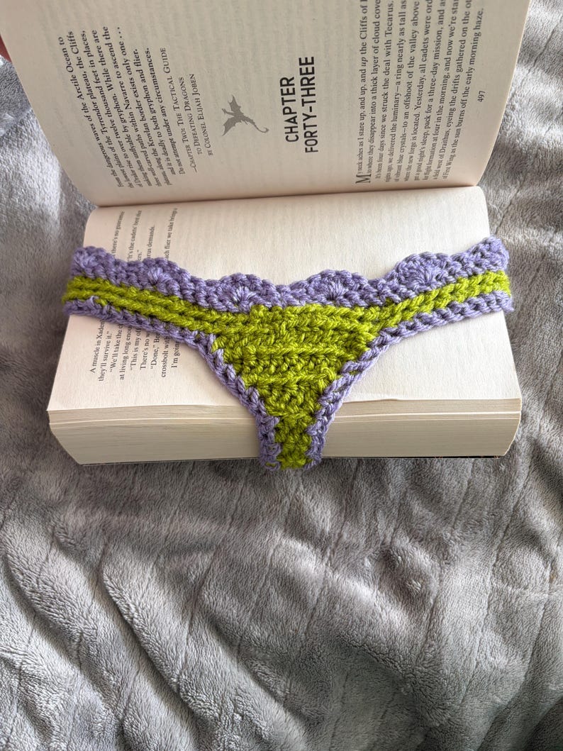 May include: A handmade crochet bookmark featuring a triangular lime green center and a scalloped lavender purple border. The bookmark is draped over the open pages of a book, with the text Chapter Forty-Three visible on the page above it.
