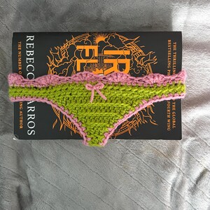 May include: A handmade crochet bikini bottom in lime green with a pink scalloped trim and a small pink bow at the center. The item is placed on top of a black book titled Iron Flame by Rebecca Yarros, which features orange graphic details.