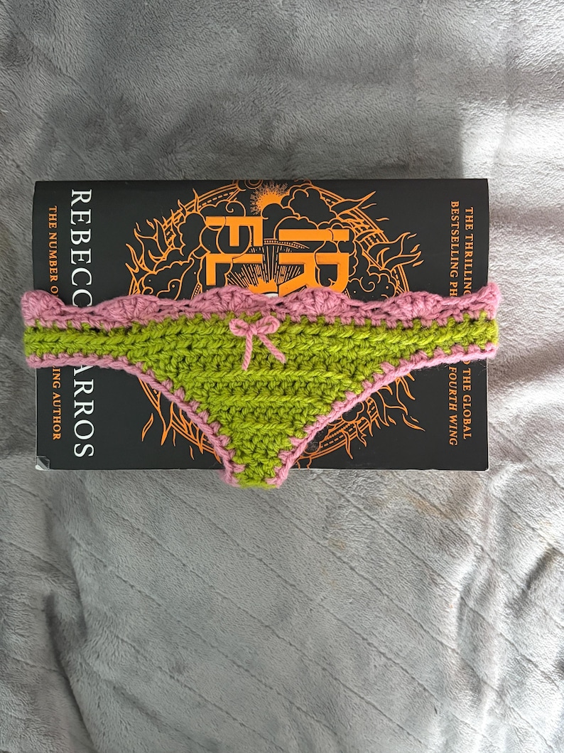 May include: A handmade crochet bikini bottom in lime green with a pink scalloped trim and a small pink bow at the center. The item is placed on top of a black book titled Iron Flame by Rebecca Yarros, which features orange graphic details.