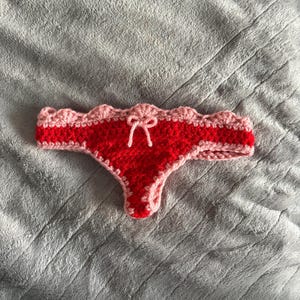 May include: A crochet thong featuring a vibrant red center panel and a light pink scalloped trim along the waistband and leg openings. A small light pink yarn bow is centered at the front of the waistband, adding a decorative detail to the piece.