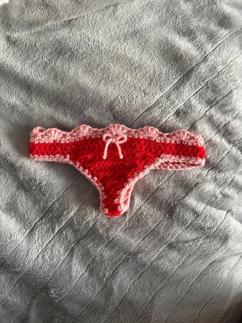 May include: A crochet thong featuring a vibrant red center panel and a light pink scalloped trim along the waistband and leg openings. A small light pink yarn bow is centered at the front of the waistband, adding a decorative detail to the piece.
