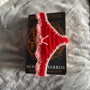 May include: A red and light pink crocheted bookmark shaped like underwear with a small bow detail. The bookmark is placed over a black book cover with the visible text Iron Flame by Rebecca Yarros, The Number One Bestselling Author.