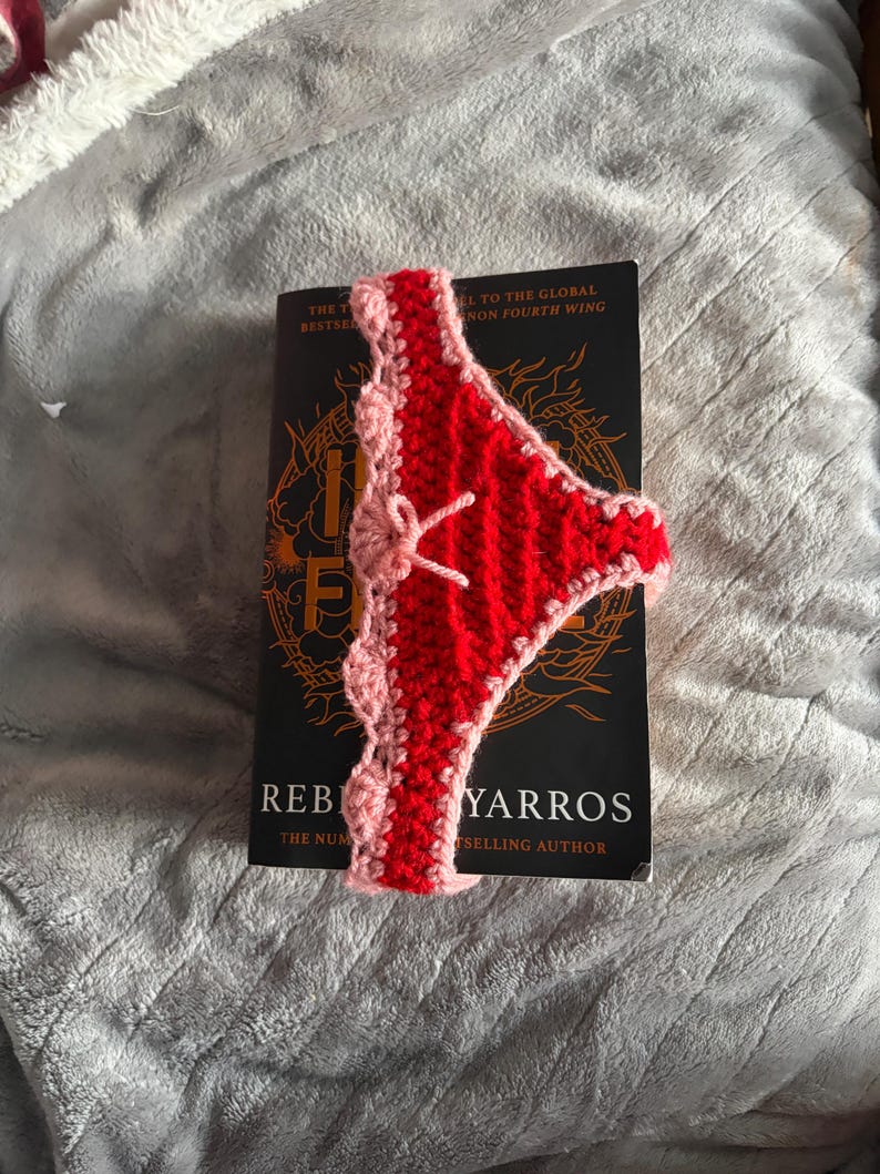 May include: A red and light pink crocheted bookmark shaped like underwear with a small bow detail. The bookmark is placed over a black book cover with the visible text Iron Flame by Rebecca Yarros, The Number One Bestselling Author.