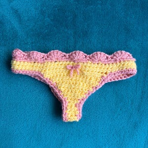 May include: A pair of crochet underwear featuring a bright yellow center panel and a scalloped pink border along the waistband and leg openings. A small pink yarn bow is centered at the top of the yellow section, creating a textured finish.