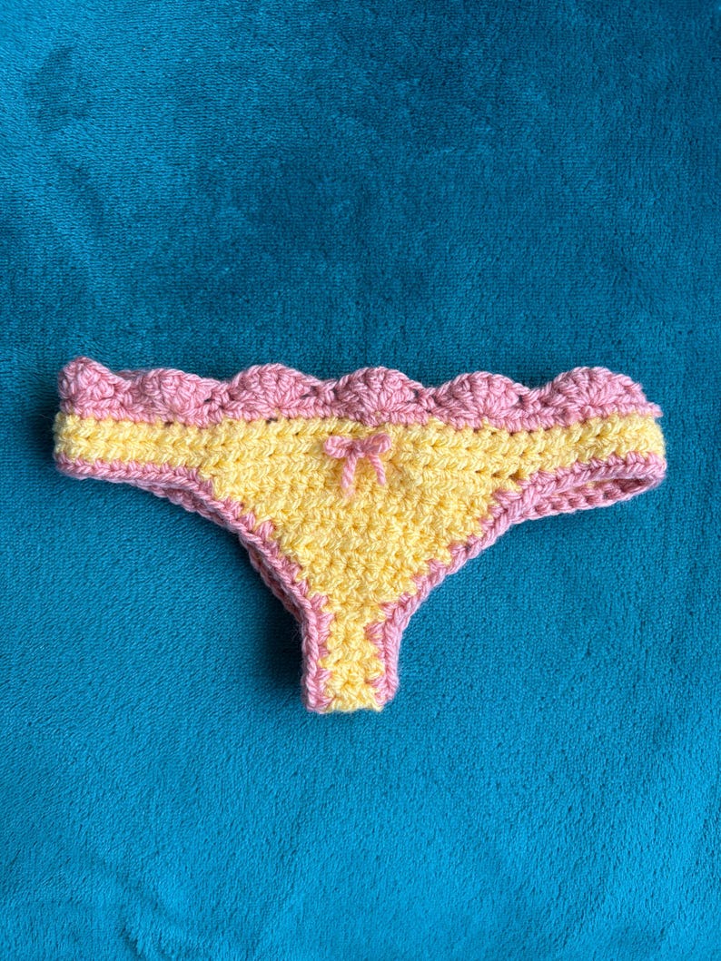 May include: A pair of crochet underwear featuring a bright yellow center panel and a scalloped pink border along the waistband and leg openings. A small pink yarn bow is centered at the top of the yellow section, creating a textured finish.