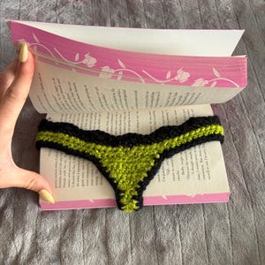 May include: A crochet bookmark shaped like a thong, featuring a vibrant lime green center panel bordered by a thick black yarn trim with a scalloped edge, placed between the pages of an open paperback book with pink floral cover accents.