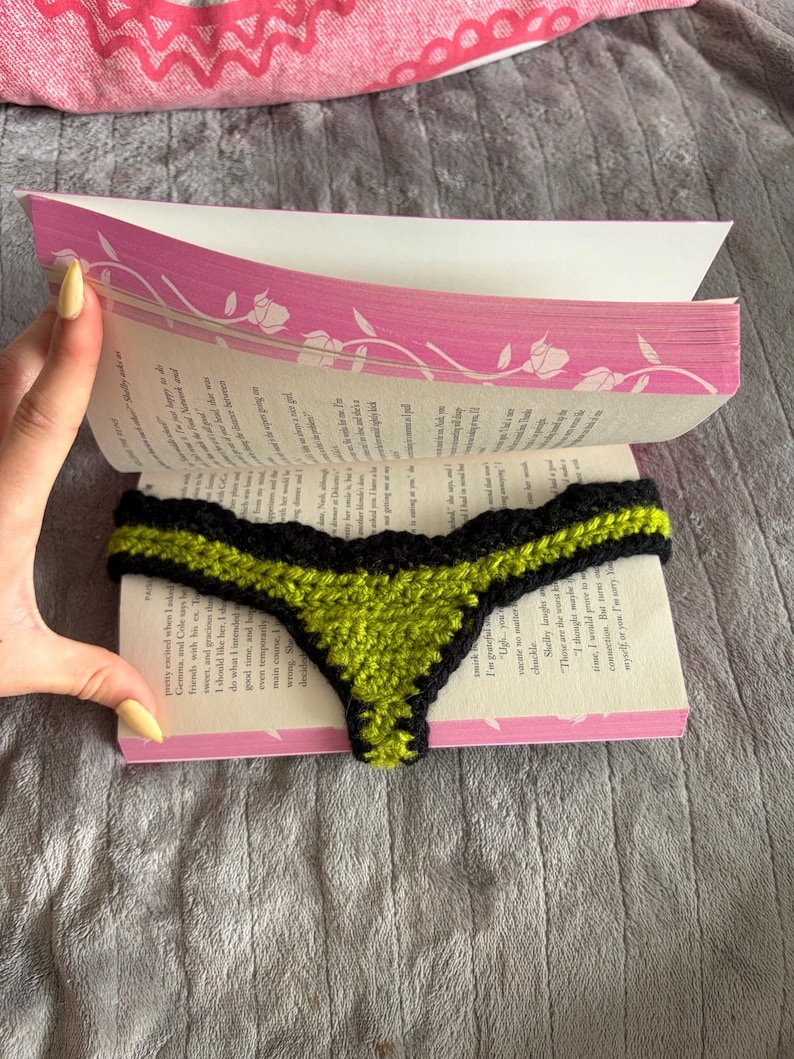 May include: A crochet bookmark shaped like a thong, featuring a vibrant lime green center panel bordered by a thick black yarn trim with a scalloped edge, placed between the pages of an open paperback book with pink floral cover accents.