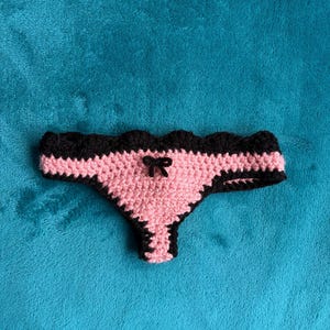 May include: A crochet thong featuring a light pink center panel with a small black bow at the center, bordered by a scalloped black trim along the waistband and leg openings, crafted with a textured yarn stitch pattern throughout the garment.